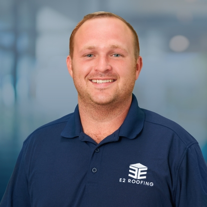 Kaleb Carson, Production & Logistics Manager at E2 Roofing, smiling in a navy polo shirt with the company logo, against a blurred background.